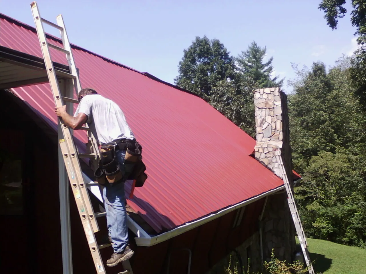 Roofer on a ladder beside a red metal roof and stone chimney — Soffit & Fascia Repair by Roofing Brothers in Onalaska